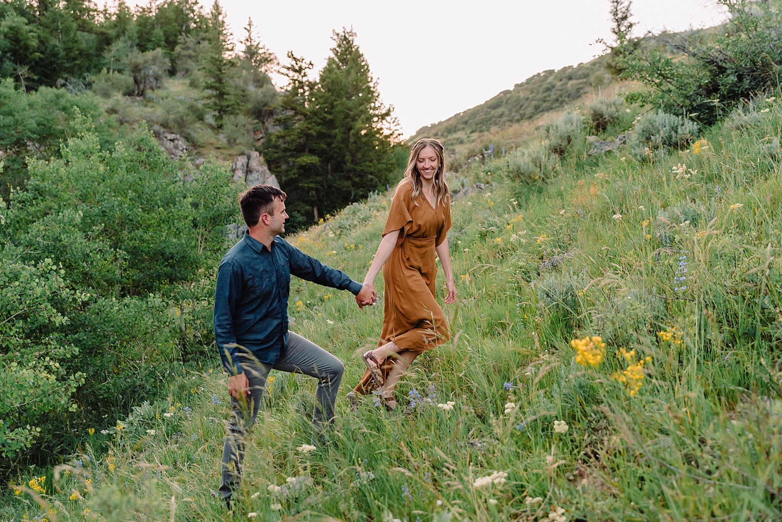 engaged couple walking up a hill while woman is wearing rust colored long dress and man wears coordinating navy shirt