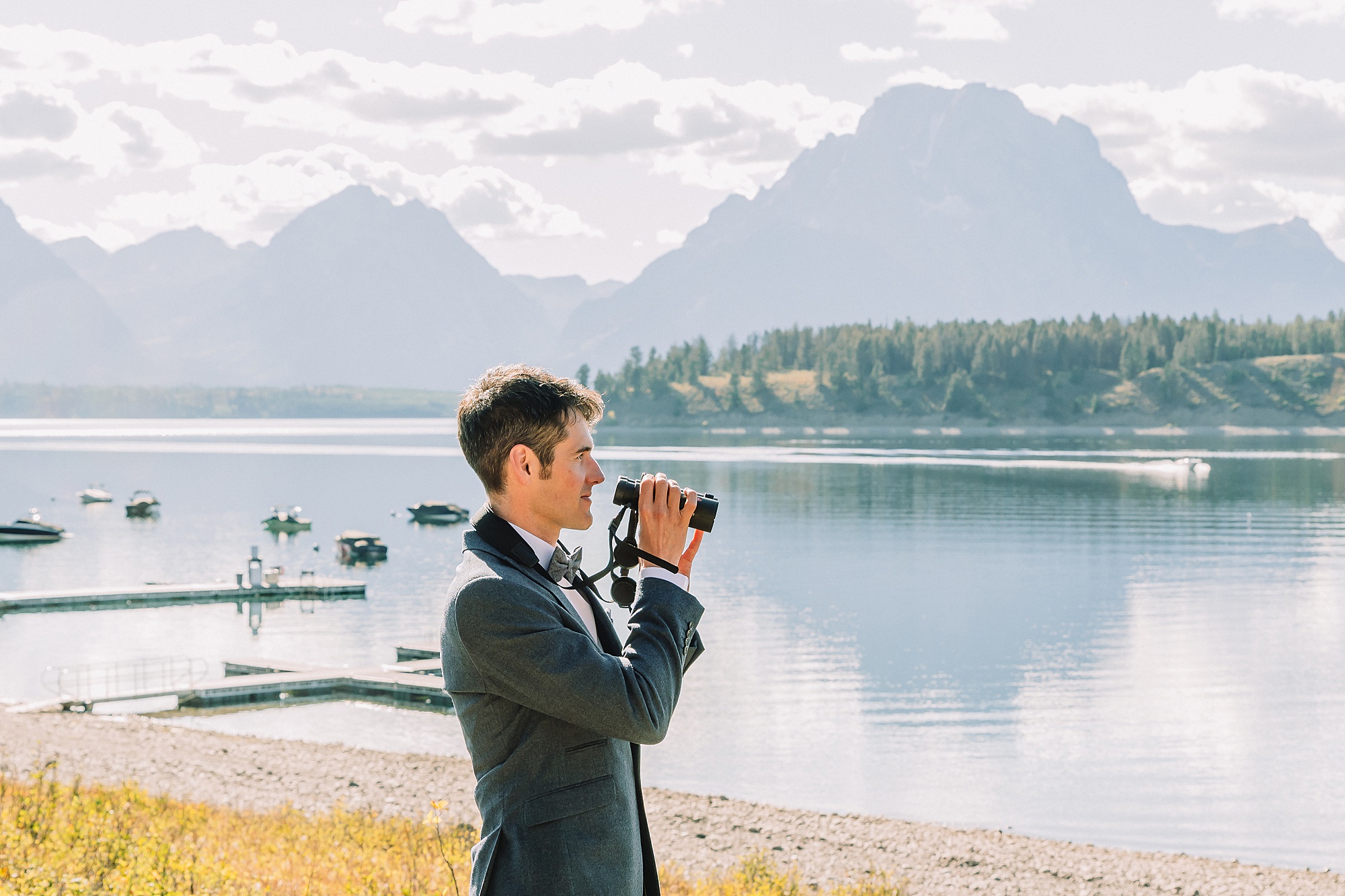 groom poses with hobby binoculars for a photo at signal mountain lodge with Jackson Lake in the background