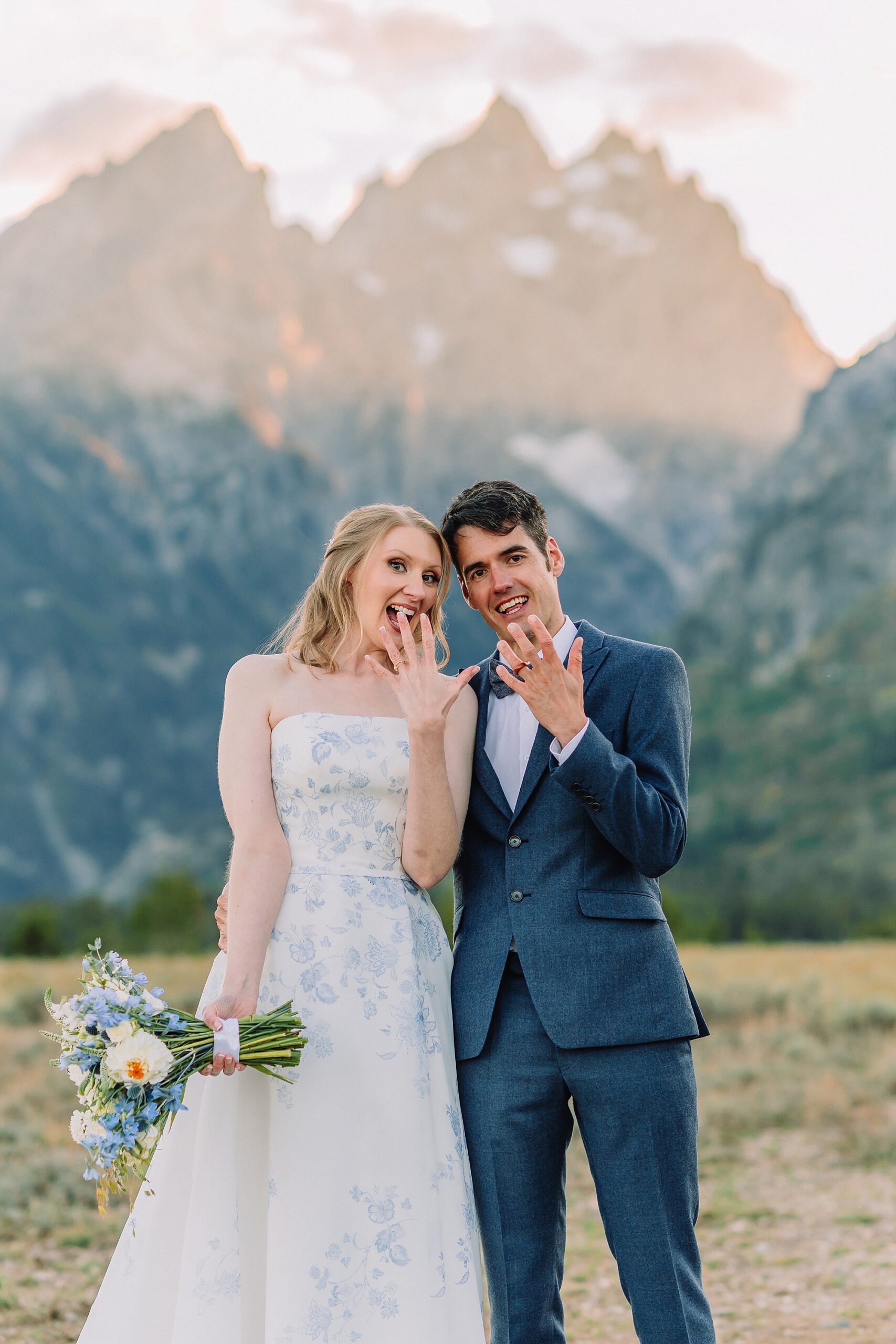 couple shows off wedding rings while wearing wedding attire in front of the Teton mountains