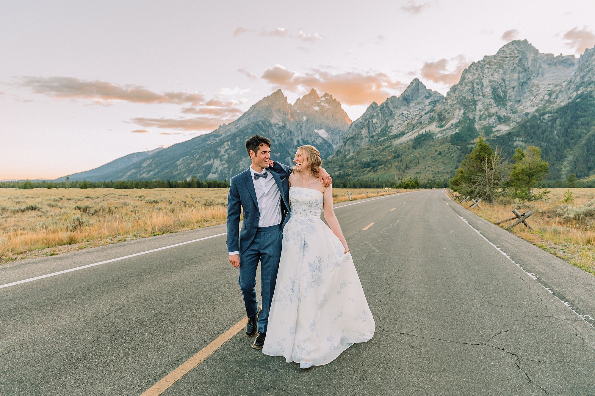 couple walks in middle of an empty road and laughs with the Cathedral Group of Grand Teton National Park behind them