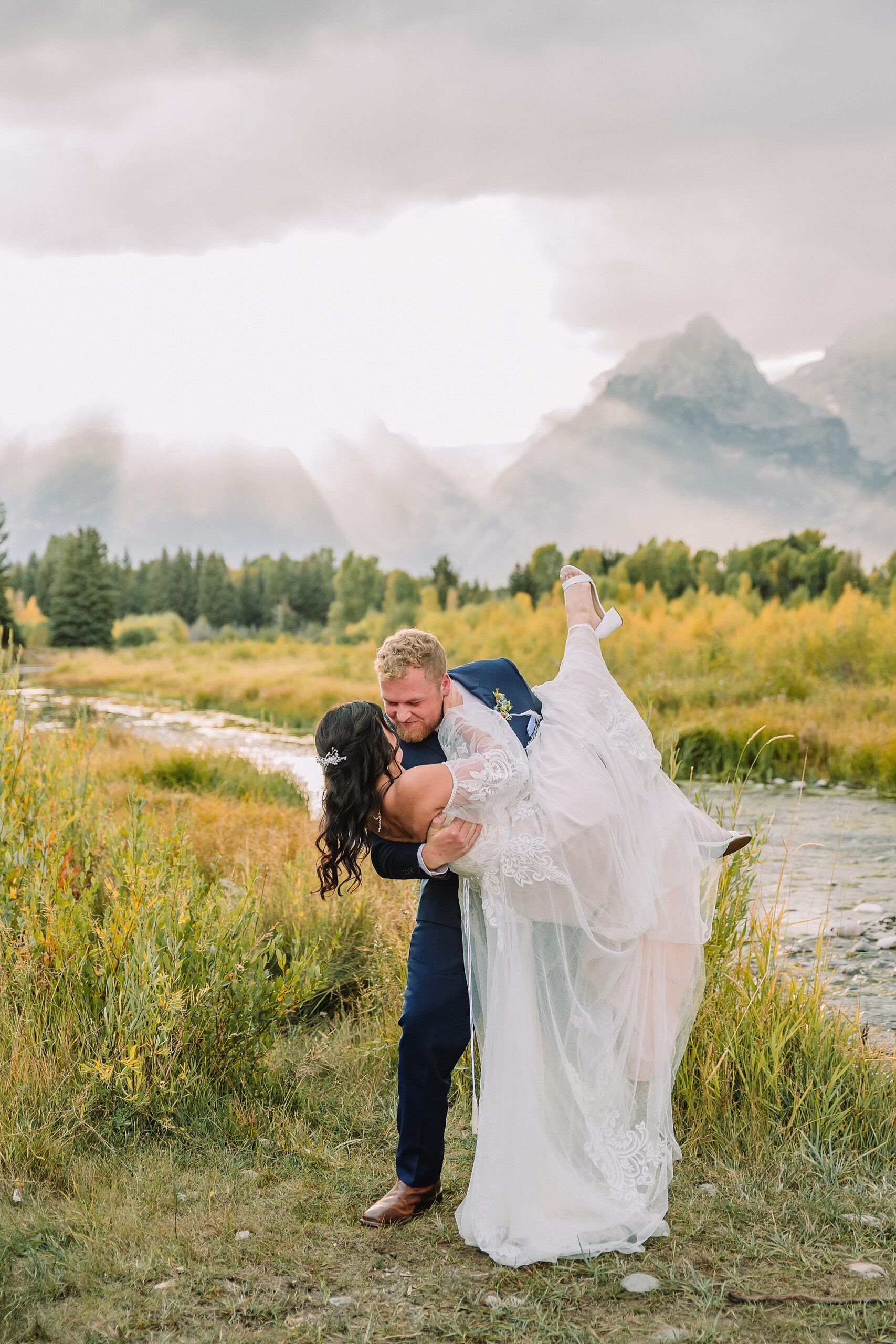groom picks up bride in romantic lift pose in front of the Teton mountains at Schwabacher Landing with God Rays behind them.