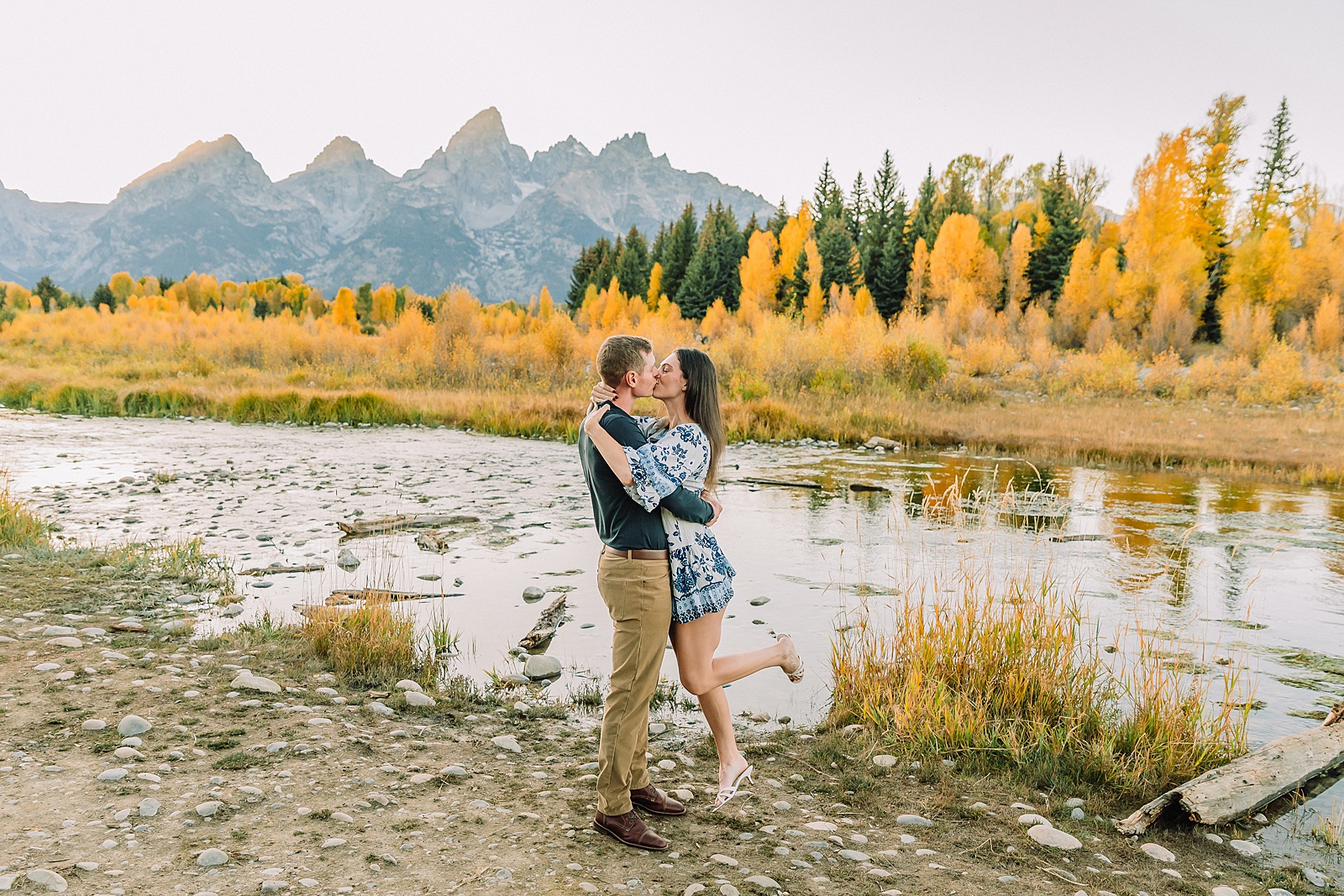 Jackson Hole engagement photos at Schwabacher Landing fall colors couple portraits along Snake River Wyoming autumn surprise engagement session Grand Teton National Park western hats engagement photos Jackson Wyoming fall engagement photos sagebrush Tetons backdrop blue dress engagement photos Jackson Hole Wyoming Schwabacher Landing couples photography fall season golden cottonwood trees engagement portraits Wyoming