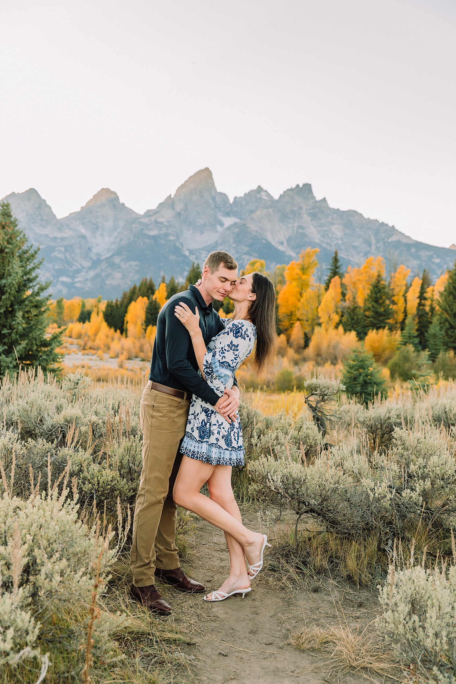 Jackson Hole engagement photos at Schwabacher Landing fall colors couple portraits along Snake River Wyoming autumn surprise engagement session Grand Teton National Park western hats engagement photos Jackson Wyoming fall engagement photos sagebrush Tetons backdrop blue dress engagement photos Jackson Hole Wyoming Schwabacher Landing couples photography fall season golden cottonwood trees engagement portraits Wyoming