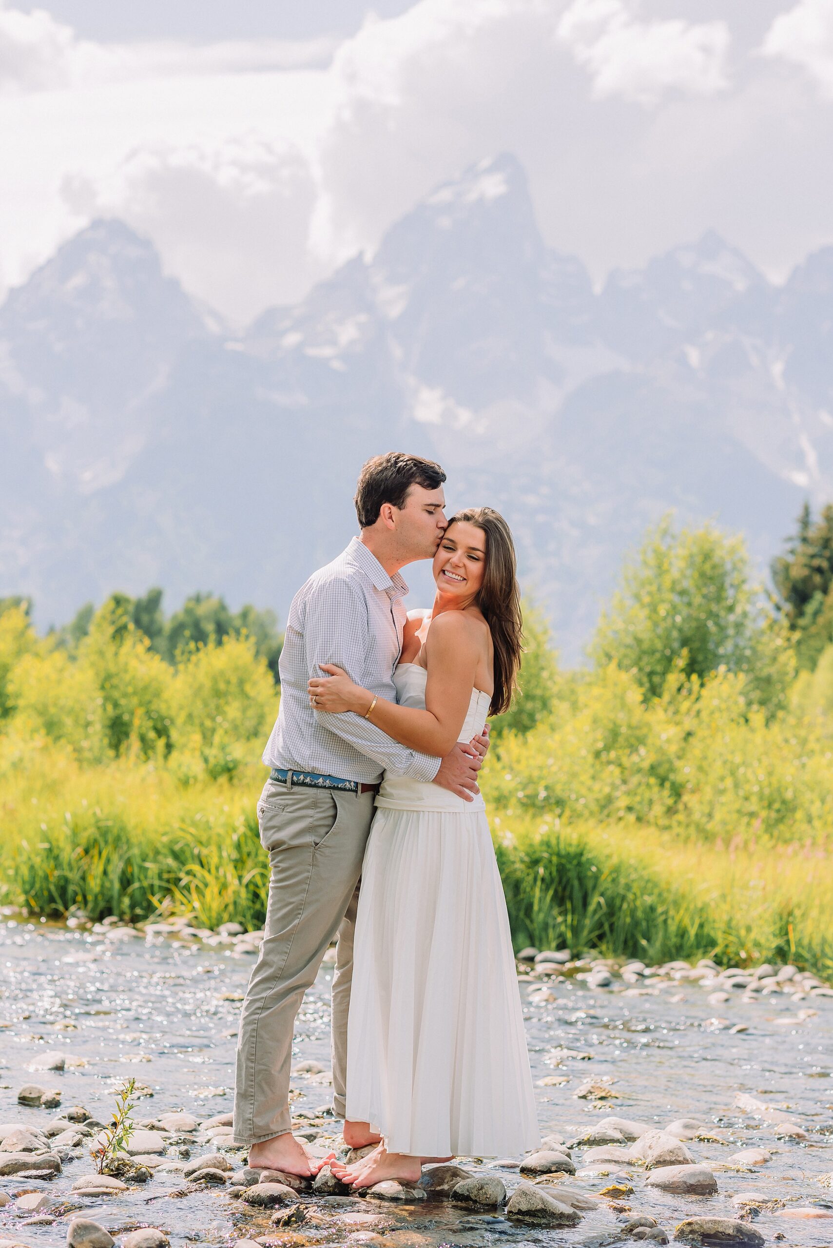 summer engagement photos at Schwabacher Landing Wyoming couple wading in river during engagement photoshoot Teton mountain backdrop engagement photography romantic nature engagement session July Wyoming outdoor engagement photos with cloudy blue skies