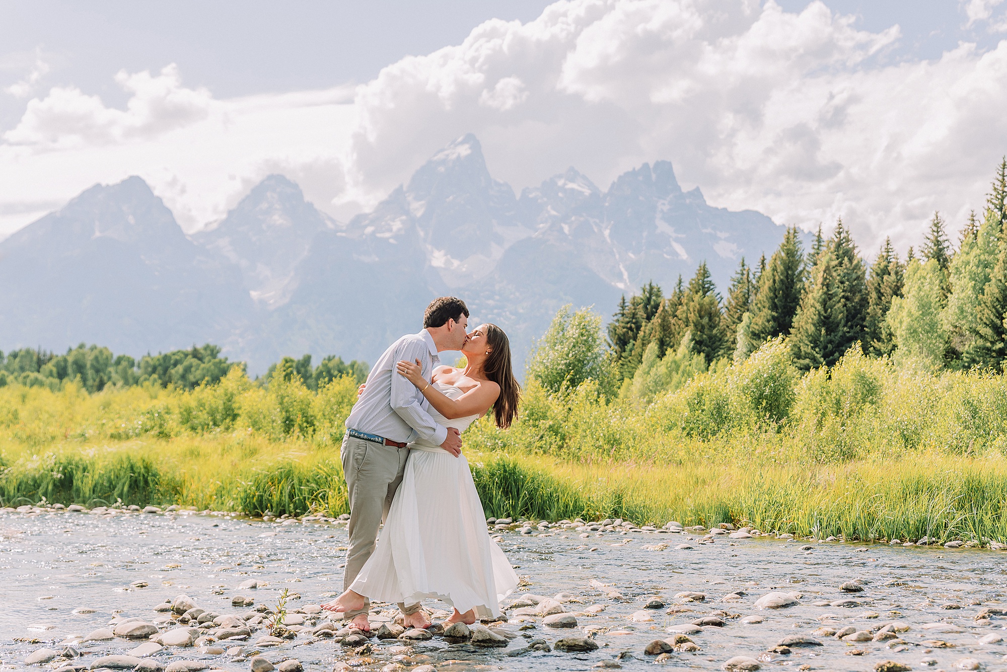 summer engagement photos at Schwabacher Landing Wyoming couple wading in river during engagement photoshoot Teton mountain backdrop engagement photography romantic nature engagement session July Wyoming outdoor engagement photos with cloudy blue skies