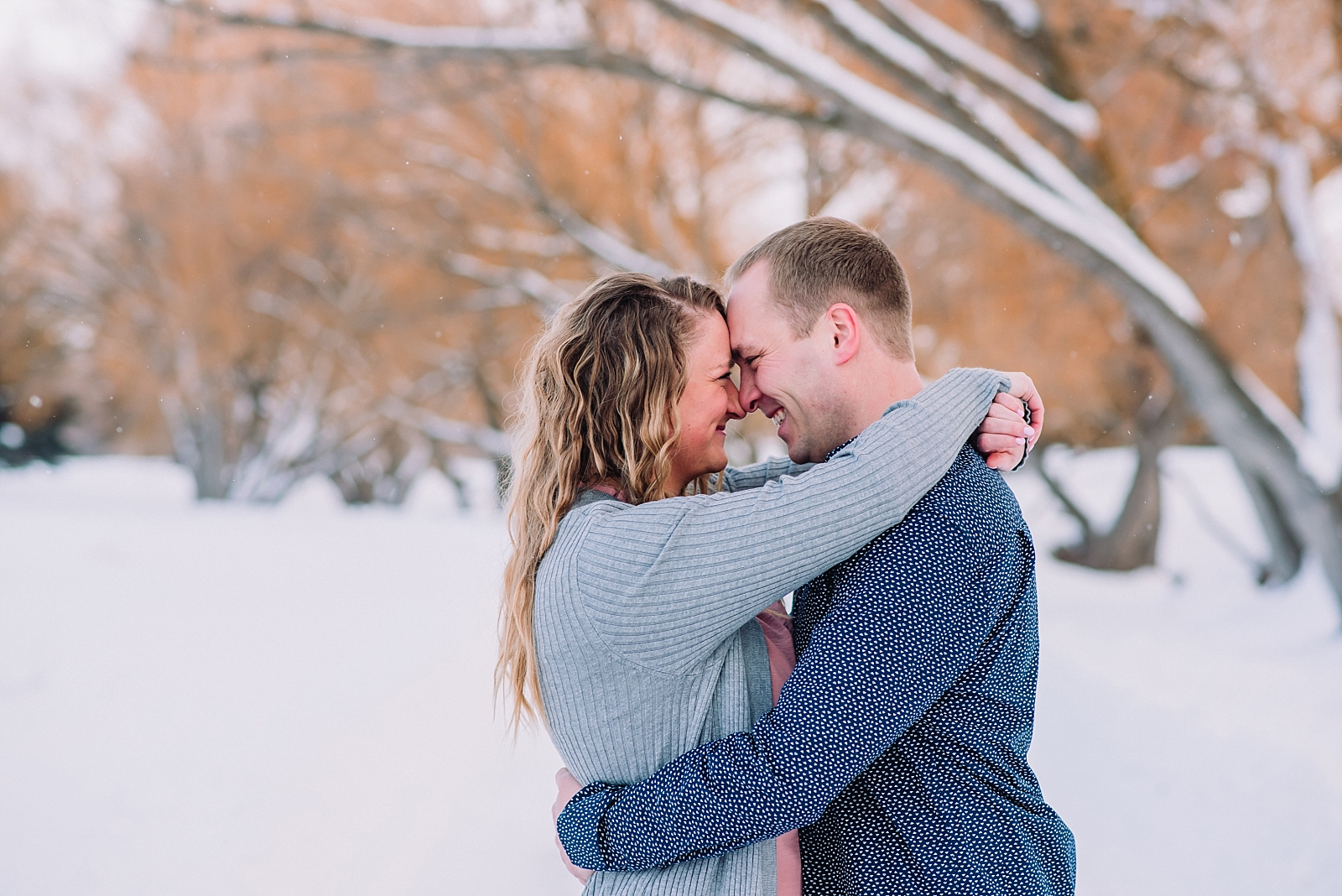 engaged couple hugging and cuddling on snowy bank with trees winter photos wedding photographer idaho falls freeman park