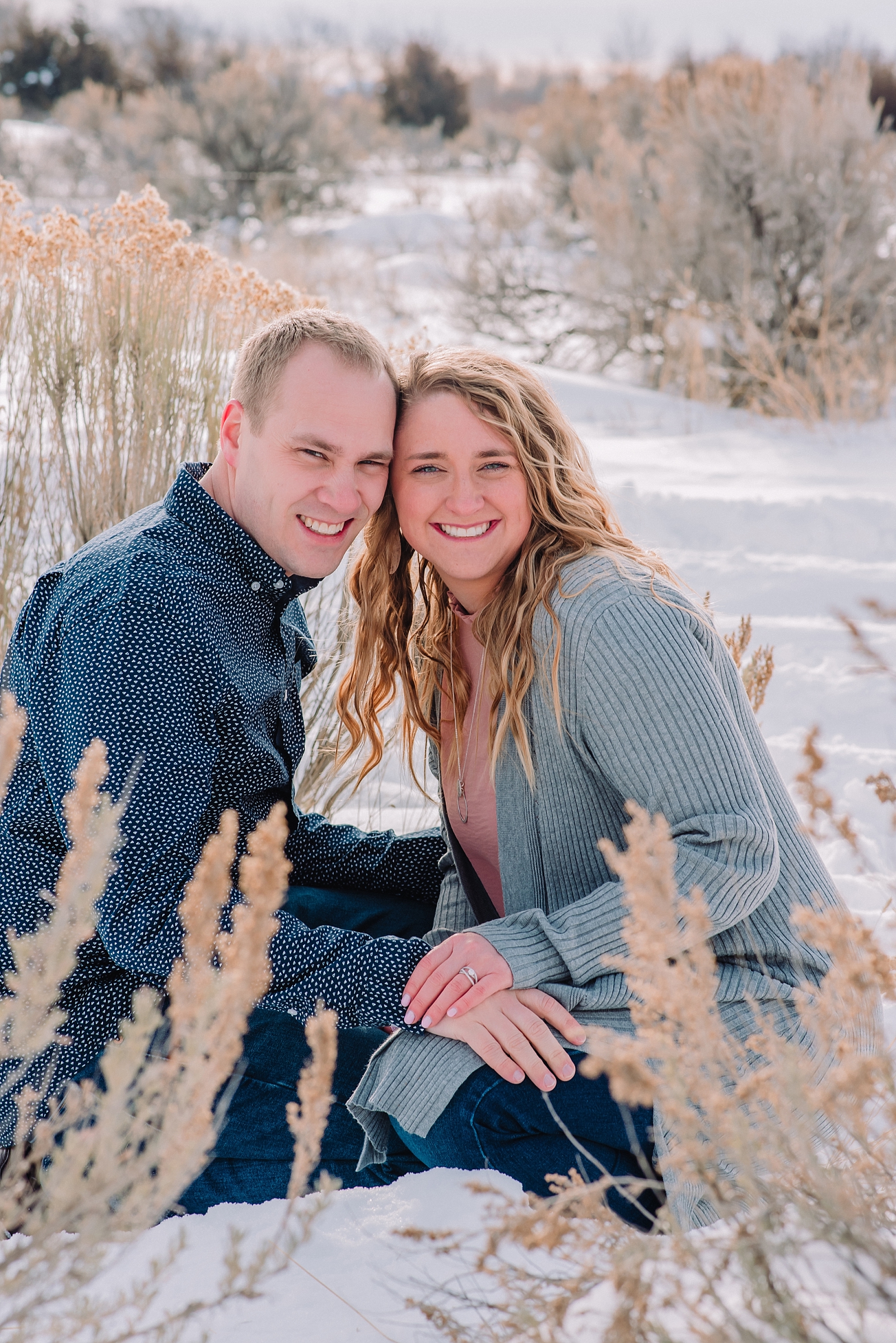 rexburg engagement photographer janelle and co engaged couple holding hands and sitting in the snow.