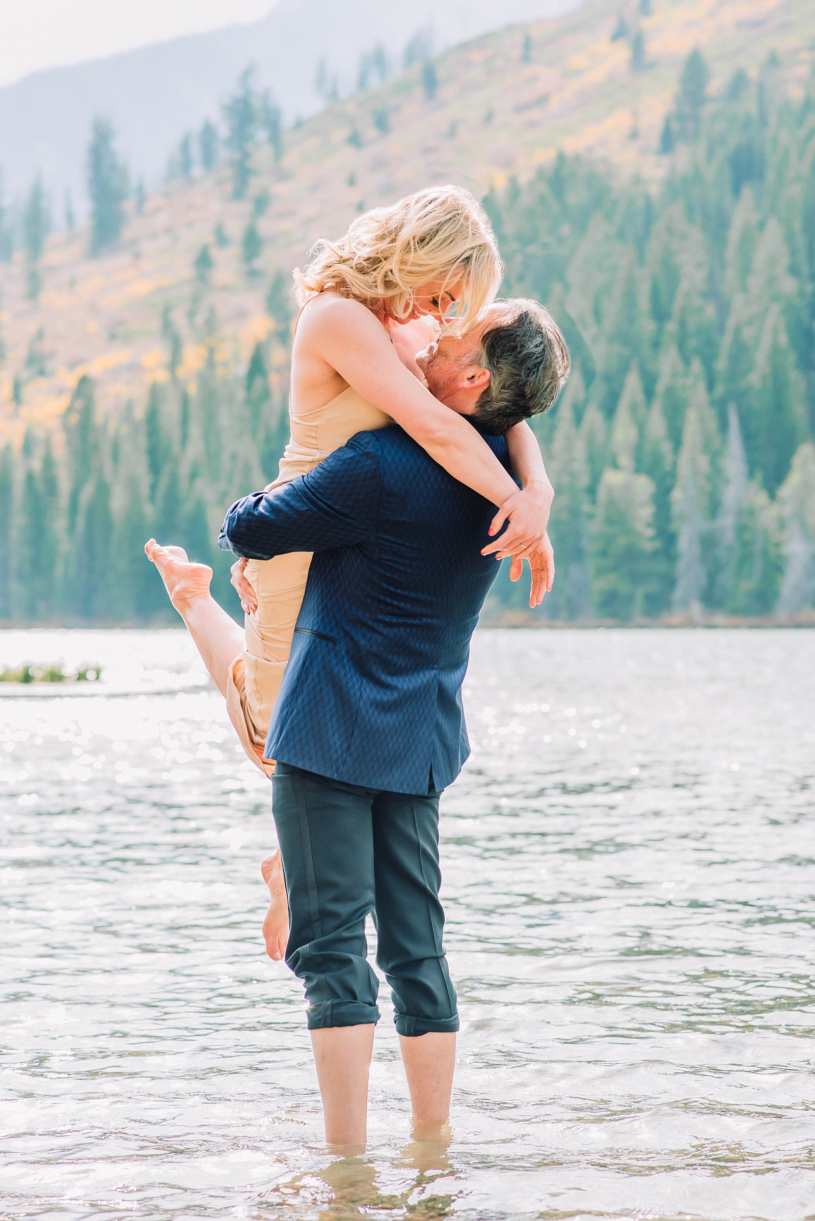 groom picks up his bride as he stands in a lake and she looks down at him and smiling