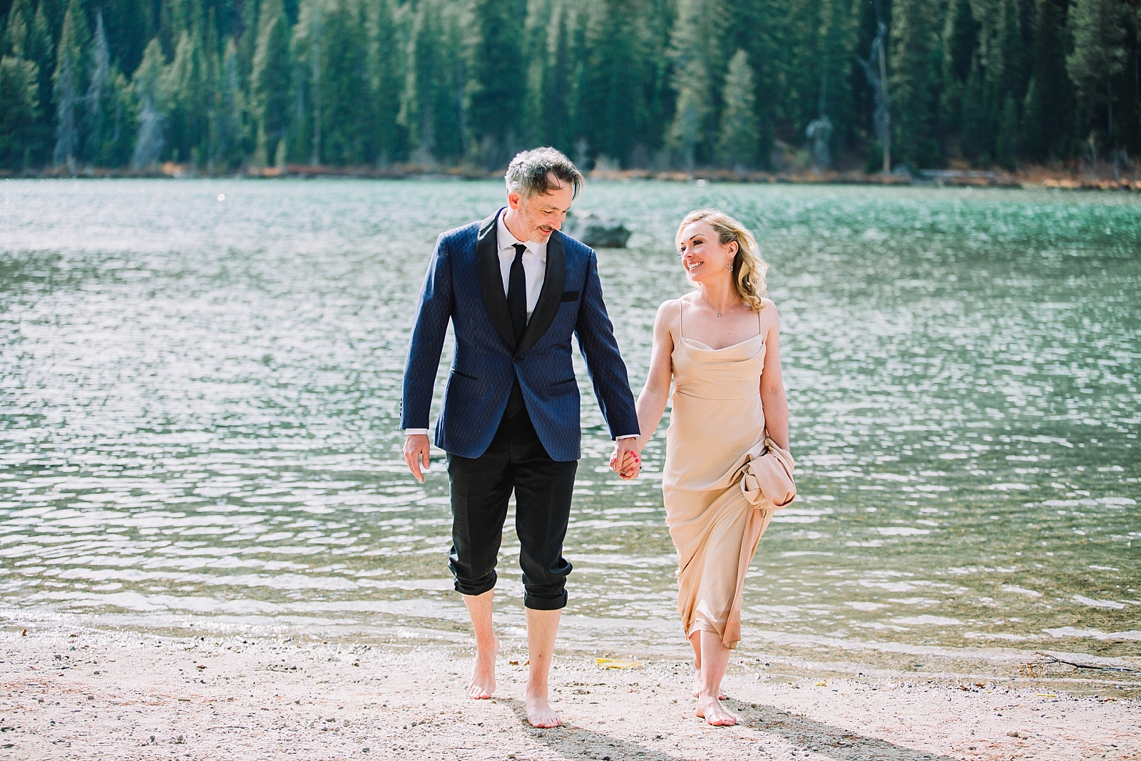 bride and groom hold hands as they walk towards the camera and away from a bright blue lake in the Tetons