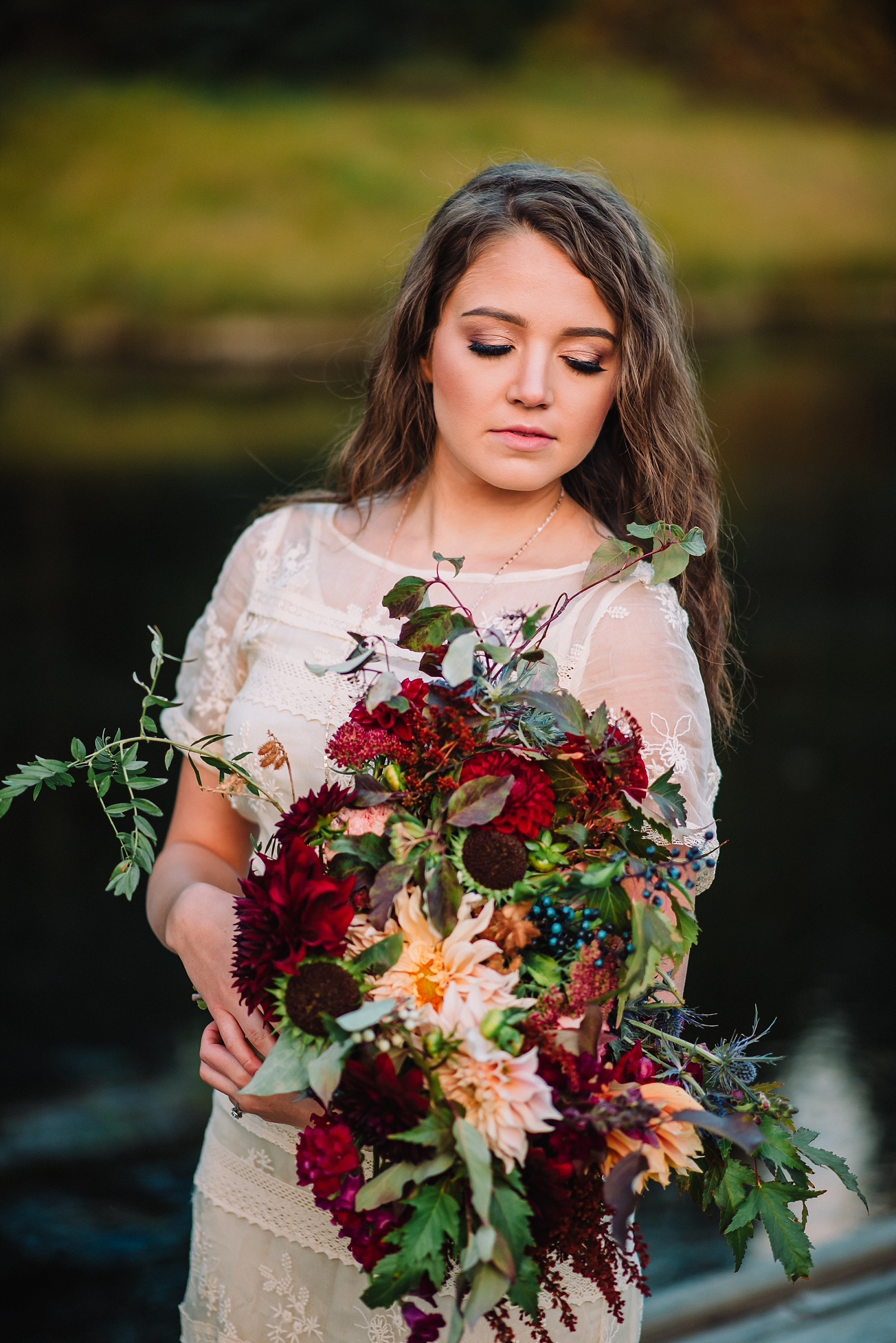 Island Park Elopement with bride holding an earht fall bouquet in front of a lake wearing a lace boho wedding gown
