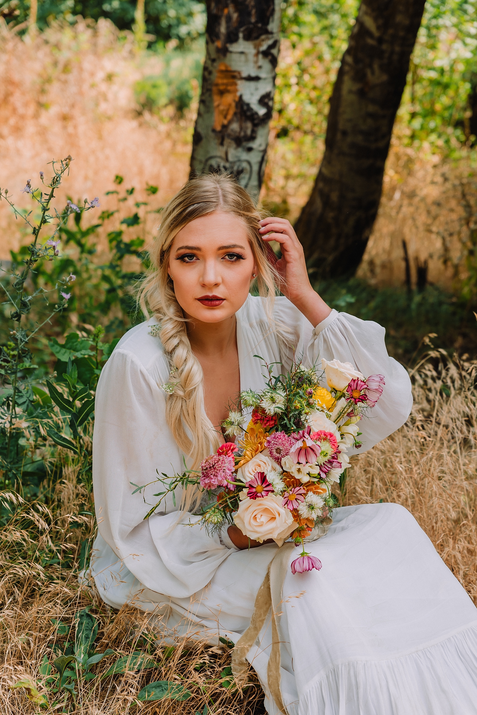 Bride sitting on ground in kelly canyon brushing hair away holding colorful bouquet
