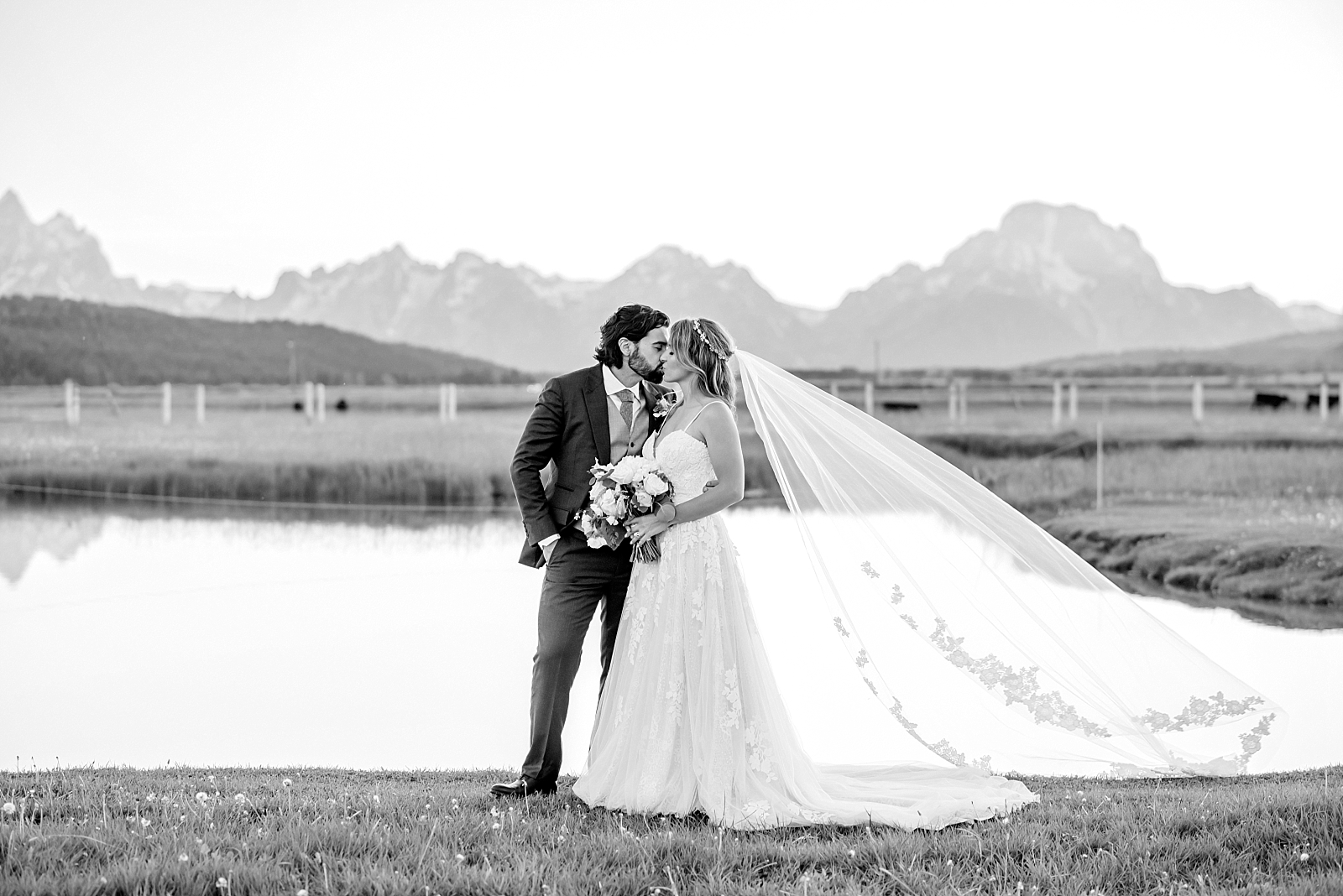 black and white image of bride and groom kissing next to a lake with the Tetons behind them 
