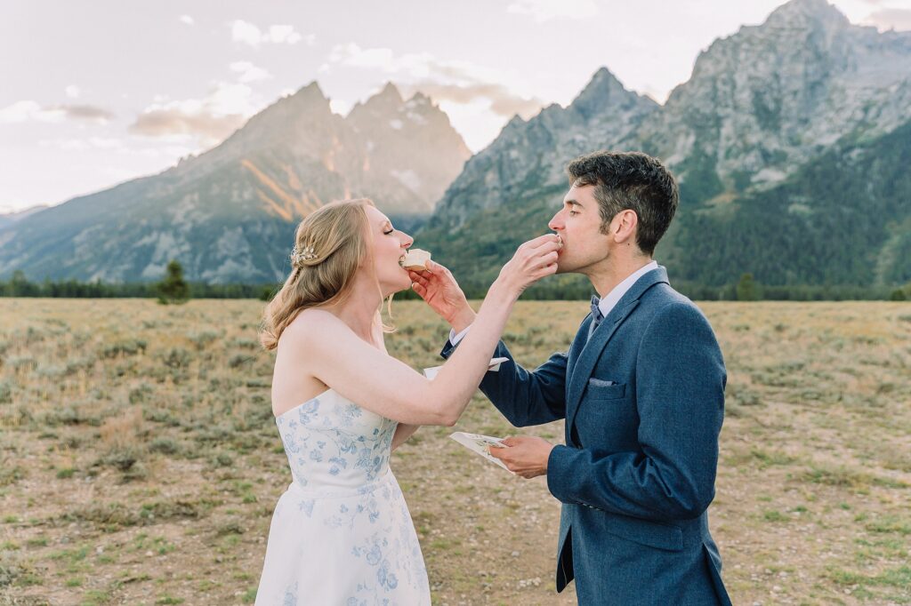 couple eats cake in wedding attire in front of the Teton mountains