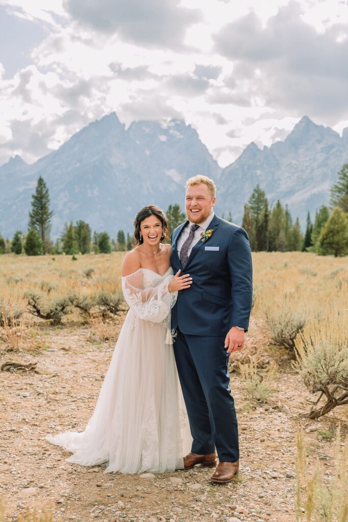 bride and groom pose and laugh in front of the Teton mountains at Mountain View Turnout in Grand Teton National Park