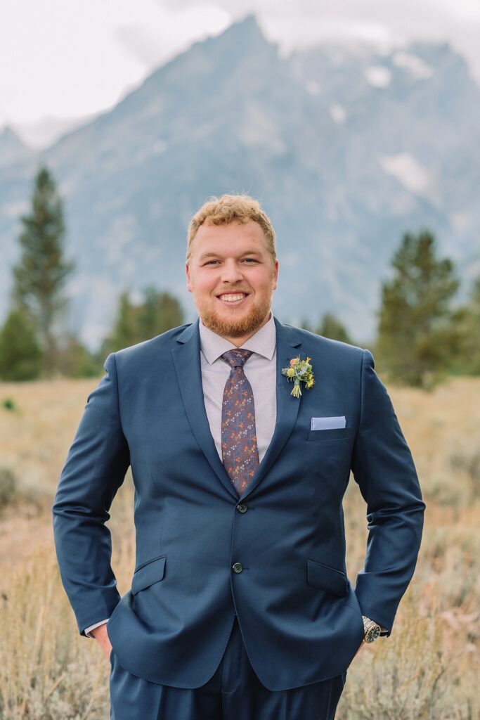 groom poses in front of mountains in blue suit and boutonniere