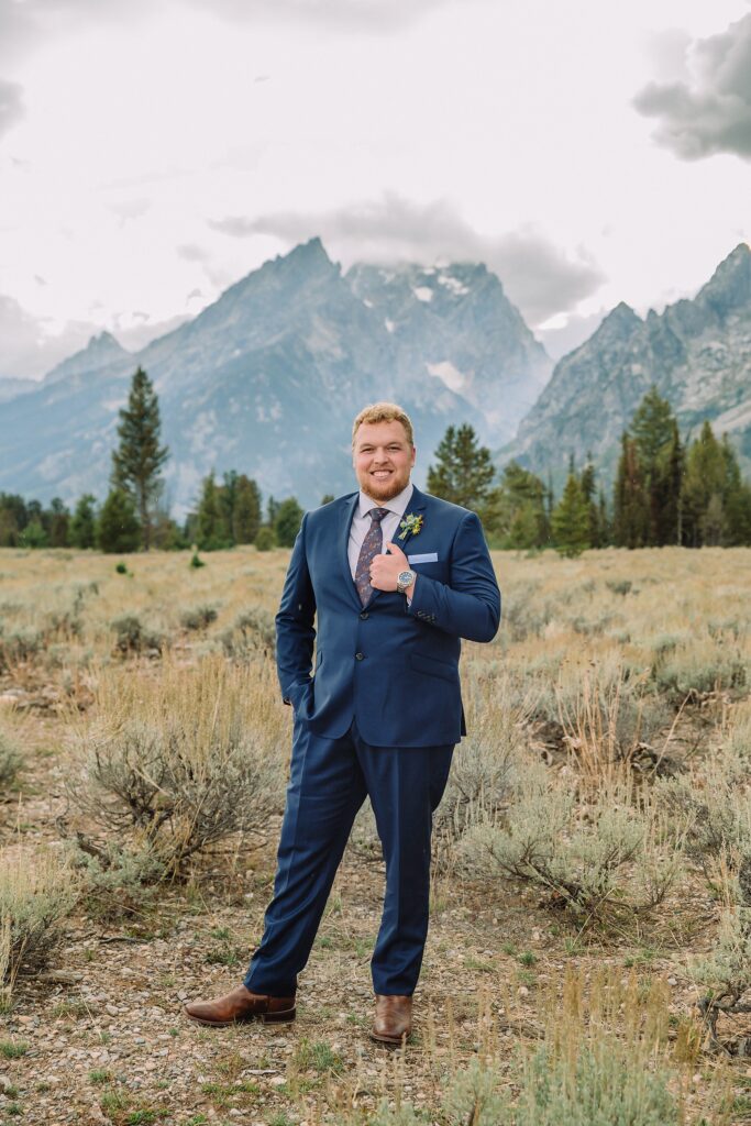 groom poses in front of mountains in blue suit and boutonniere